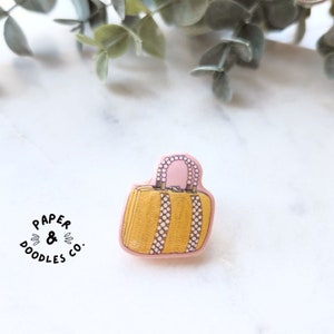 Op de afbeelding: Een gele en roze emaille pin met een cartoonillustratie van een handtas met een parelmoerachtige rand. De pin heeft de tekst "PAPER & DOODLES CO." gedrukt onder de handtas.