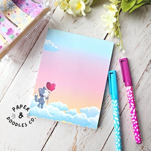 May include: A pastel pink and blue notepad featuring a cartoon cat holding two red heart balloons. The cat is perched on a cloud against a backdrop of a blue sky and white clouds. The notepad has the text "PAPER & DOODLES CO." printed in the bottom left corner.