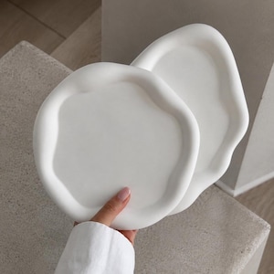 May include: Two matte white trays with irregular, organic, wavy edges. The trays feature a smooth, solid surface with a raised rim and a shallow basin, designed with a non-uniform, rounded shape that lacks sharp corners or straight lines.