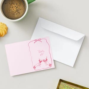 May include: A pink greeting card with a bow design and the words "Twenty ate" in a handwritten font. The card is next to a white envelope, a green mug of coffee, and two cookies.