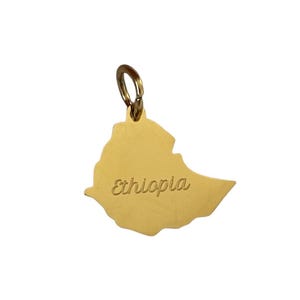 Puede incluir: Encanto de metal dorado en forma del contorno de Etiopía con la palabra "Etiopía" inscrita en el encanto.