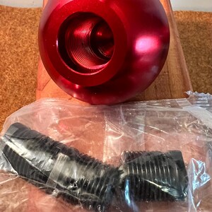 May include: A bright red, spherical metal component featuring a large, threaded circular opening at its center. Below it, two black, cylindrical, threaded plastic adapters are enclosed within a clear, crinkled plastic storage bag.