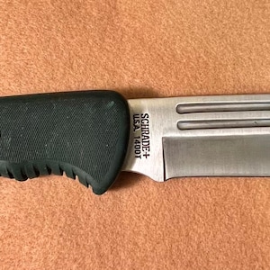 May include: A stainless steel knife with a curved blade and a dark green handle. The handle is labeled "OLD TIMER". The blade has the text "SCHRADE+ USA 140OT". The knife is lying on a brown surface.