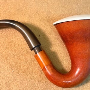 May include: A tobacco pipe featuring a smooth, reddish-brown wooden bowl with a wide, flared top and a white lid. The pipe has a curved, dark brown stem connected to the bowl by a translucent amber-colored band, creating a distinct, functional shape.