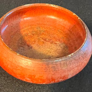 May include: A round, shallow bowl with a vibrant orange exterior and interior. The surface features a textured, mottled finish with visible signs of wear along the upper rim, revealing lighter tones beneath the primary orange color.