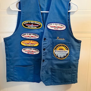 May include: A bright blue vest featuring multiple embroidered patches from the Academy of Model Aeronautics, including Instructor, Pilot, Radio Control, Mechanic, and All Season Flyer. The name Ross is embroidered in yellow thread above the chest pocket.