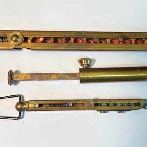 May include: Three antique brass tools are displayed against a white background. The top tool is a long, rectangular piece with red cylindrical objects visible inside. The middle tool is cylindrical with a knob. The bottom tool has a black handle.