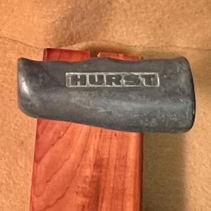 May include: A dark gray sledgehammer head with the word "HURST" in white lettering is mounted on a reddish-brown wooden handle. The tool is designed for heavy-duty tasks and construction.