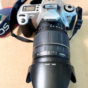 May include: A silver Canon Rebel EOS 2000D camera with a black Tamron lens. The lens has measurement markings. The camera strap is blue with white "Canon" text. The camera is on a beige surface.