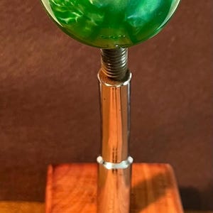 May include: A custom shift knob with a green and white swirl design. The knob is mounted on a chrome shaft and sits on a wooden base. Three brass threaded inserts are also included.
