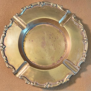 May include: A round brass ashtray featuring a smooth central basin and a wide, decorative rim with four integrated notches. The metal surface displays a warm, metallic gold tone with visible signs of natural oxidation and patina throughout.