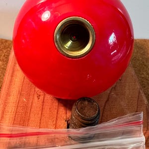 May include: A glossy, spherical red object featuring a central circular brass-colored metal insert with a threaded opening. Positioned below the sphere is a small, cylindrical, dark-colored metal component with visible threading and a clear plastic bag.