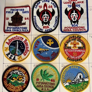 May include: A collection of vintage Boy Scout patches. The patches feature various designs, including a rocket, a Native American figure, and text such as "Frontier Days" and "Camp Long Lake." The patches are embroidered with vibrant colors and text.