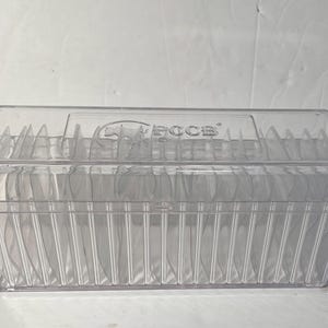 May include: A clear, rectangular plastic container with a hinged lid. The container is filled with multiple clear, oval-shaped objects. The lid has the text "PCCB" printed on it. The container is on a white surface.