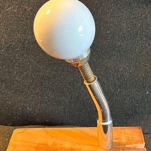 May include: A white spherical knob attached to a curved, metallic, silver-toned arm. The arm features a threaded section near the base of the sphere and connects to a smooth, cylindrical metal post that is mounted onto a light-colored wooden base.