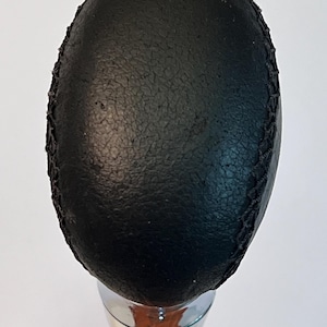 May include: A black, oval-shaped gear shift knob with a textured surface and stitched edging. The knob is mounted on a shiny, chrome-colored metal shaft. The background is plain white.