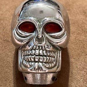 May include: A chrome skull-shaped gear shift knob with red eye sockets. The skull has detailed teeth and facial features. The metal is shiny and reflective, with a few minor imperfections. The knob is designed to be attached to a gear shift lever.
