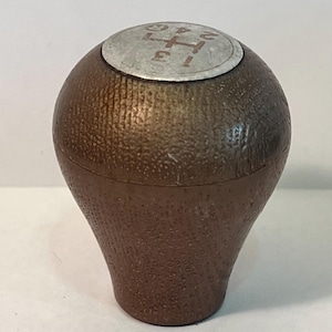 May include: A brown gear shift knob with a textured surface. The top of the knob is silver with a red gear pattern and numbers 1, 2, 3, 4, and R. The knob has a rounded top and a tapered base.