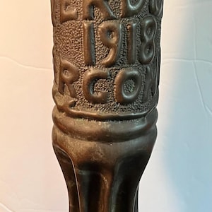 May include: A tall, cylindrical bronze-colored metal object with a textured surface featuring embossed lettering that reads NOV, VERDUN, 1918, RCON. The object has a flared, fluted base and a smooth, rounded top edge, showing a dark, weathered patina.