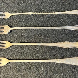 May include: Four silver-toned metal cocktail forks arranged horizontally. The top fork has a twisted handle and two tines. The second, third, and fourth forks have three tines and varying decorative patterns on their handles, including embossed details.