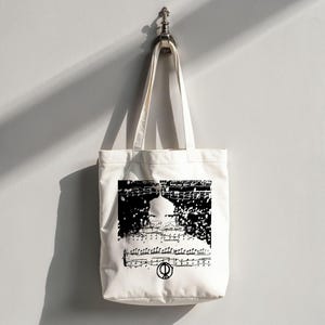 May include: Off-white tote bag with a black and white musical score design. The design includes musical notes and a symbol. The bag has long straps for carrying over the shoulder.
