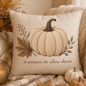May include: A beige decorative pillow with an embroidered pumpkin design. The pumpkin is light beige with a brown stem, surrounded by brown leaves and sprigs. The text "a season to slow down" is below the pumpkin. A lit candle and small pumpkins are in the foreground.