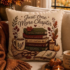 May include: A decorative pillow with the text "Just One More Chapter" and a stack of books, a mug of coffee, and a candle. The pillow has a fall theme with autumn colors and imagery. A cozy reading scene.