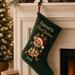 Op de afbeelding: Een donkergroene kerstsok met een geborduurde illustratie van een rendier met een rode kerstmuts en een groene strik. De tekst luidt Daniel's First Christmas en 2026, geaccentueerd met gouden geborduurde sneeuwvlokken.