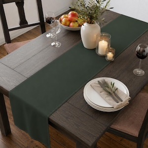 Puede incluir: Un camino de mesa rectangular de color verde bosque oscuro colocado sobre una mesa de comedor de madera marr&oacute;n oscuro. El camino presenta bordes rectos cosidos y una textura de tela lisa y mate, ofreciendo una base sencilla para la mesa.