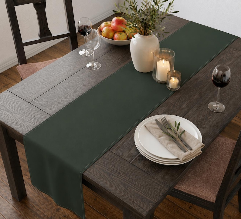 Puede incluir: Un camino de mesa rectangular de color verde bosque oscuro colocado sobre una mesa de comedor de madera marr&oacute;n oscuro. El camino presenta bordes rectos cosidos y una textura de tela lisa y mate, ofreciendo una base sencilla para la mesa.