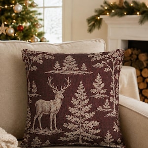 May include: A square throw pillow featuring a deep burgundy background with a woven pattern of light beige deer and evergreen trees. The design displays a repeating forest scene with detailed silhouettes of stags and various coniferous trees.