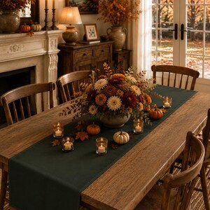 Puede incluir: Un camino de mesa rectangular de tela en color verde oscuro, colocado en el centro de una mesa de comedor de madera. El camino de mesa est&aacute; decorado con un arreglo floral central de crisantemos oto&ntilde;ales, peque&ntilde;as calabazas y velas votivas.