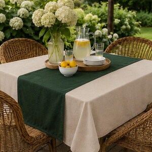 Puede incluir: Un mantel cuadrado de color beige con un camino de mesa verde oscuro, cuatro sillas de mimbre trenzado, una jarra de cristal con limonada y rodajas de lim&oacute;n, un jarr&oacute;n de cristal con hortensias blancas, un cuenco con limones y platos blancos.