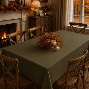 Peut inclure: Une nappe rectangulaire vert olive recouvrant une table à manger, entourée de quatre chaises en bois à dossier croisé. Un centre de table composé de citrouilles orange et blanches avec des feuilles d'automne est posé sur la table.