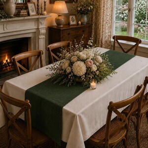 Puede incluir: Una mesa de comedor rectangular cubierta con un mantel color crema y un camino de mesa de tela verde oscuro. Un gran centro de mesa floral con flores blancas y rosas se sit&uacute;a en el centro, acompa&ntilde;ado por un peque&ntilde;o portavelas de cristal.