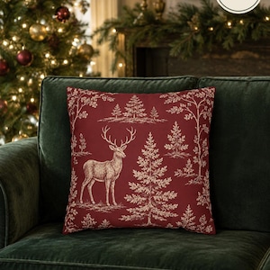 May include: A square pillow cover in deep red featuring a white line-art pattern of a standing stag and various evergreen trees. The fabric surface includes a circular white label with the text PILLOW COVER ONLY NO INSERT in black lettering.