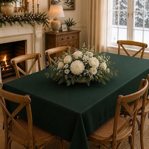May include: A rectangular dark green tablecloth draped over a dining table, surrounded by four wooden cross-back chairs. A centerpiece featuring white roses, white hydrangea, and green foliage sits in the center of the table.
