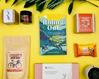 May include: A flat lay on a yellow surface features a book titled "Riding Out", a protein flapjack, a box of cyclist's hack wax, a bag of bath soak, a jar of repair balm, a box with a logo, and a selection box of coffee.