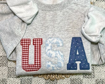 Op de afbeelding: Een grijs sweatshirt met het woord "USA" in grote, kleurrijke letters. De letters zijn gemaakt van stof met verschillende patronen: rood-witte ruit, blauwe bloemen en blauwe stippen.