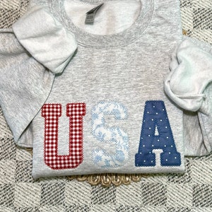 May include: A gray sweatshirt with the word "USA" spelled out in large, colorful letters. The letters are made of fabric with different patterns: red and white gingham, blue floral, and blue polka dots.