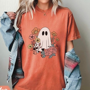 K&ouml;nnte beinhalten: Ein orangefarbenes T-Shirt mit einem wei&szlig;en Geist und einem floralen Design. Der Geist ist von orangefarbenen, gelben und rosa Blumen umgeben.