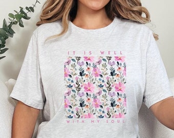 May include: A grey t-shirt with a floral print and the text "It is well with my soul" in pink.
