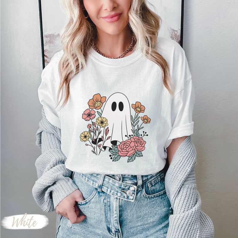 K&ouml;nnte beinhalten: Wei&szlig;es T-Shirt mit einem Aufdruck eines Geistes in einem Blumenfeld. Die Blumen sind rosa, gelb und orange.