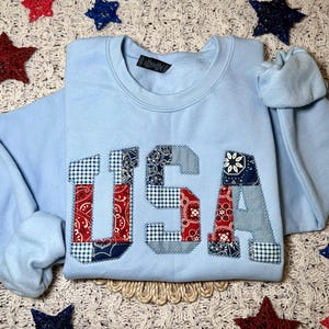 May include: A light blue crewneck sweatshirt featuring the letters USA appliqued on the front. The letters are constructed from various fabric patches in red, white, and blue, showcasing a mix of bandana, gingham, and floral patterns.
