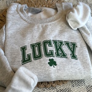 May include: A gray sweatshirt with the word "LUCKY" embroidered in green with a green four-leaf clover underneath.