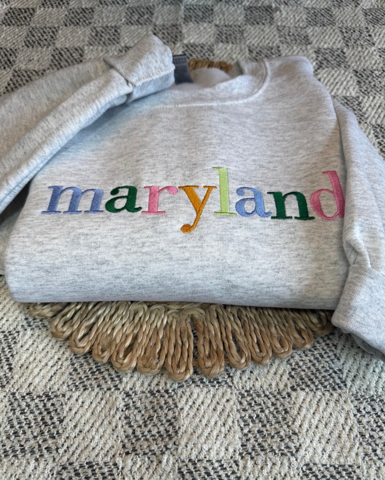 May include: A gray sweatshirt with the word "maryland" embroidered in colorful letters. The letters are in shades of blue, green, pink, orange, and yellow.