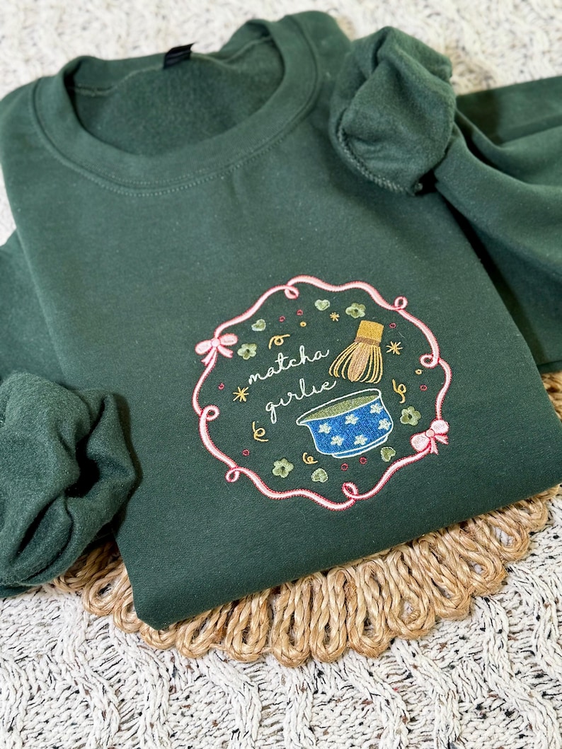 Puede incluir: Sudadera verde oscuro con un dise&ntilde;o bordado. El dise&ntilde;o presenta las palabras "matcha girlie" en un marco rosa, junto con un batidor de matcha y un cuenco azul con estrellas blancas. La sudadera est&aacute; doblada sobre una estera tejida.