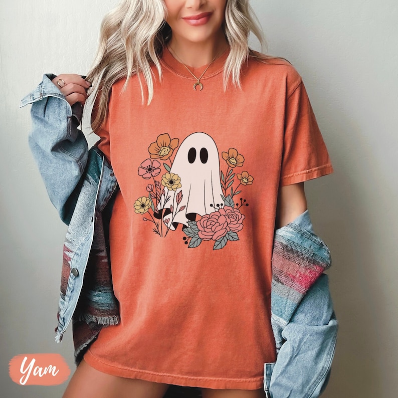 K&ouml;nnte beinhalten: Ein orangefarbenes T-Shirt mit einem wei&szlig;en Geist und einem floralen Design. Der Geist ist von orangefarbenen, gelben und rosa Blumen umgeben.