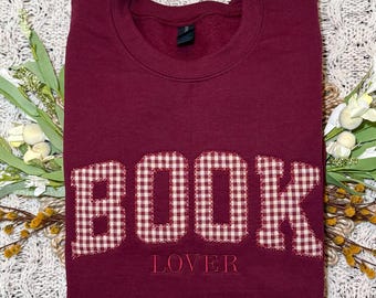 Puede incluir: Una sudadera de cuello redondo color granate que presenta la palabra BOOK en letras grandes de tela de cuadros vichy rojos y blancos con bordes deshilachados. Debajo de la palabra BOOK, la palabra LOVER está bordada en hilo rojo.