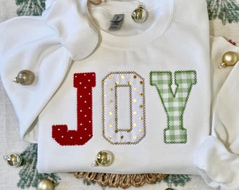 May include: White sweatshirt with the word "JOY" embroidered in red, gold, and green. The letters are decorated with polka dots and gingham patterns.